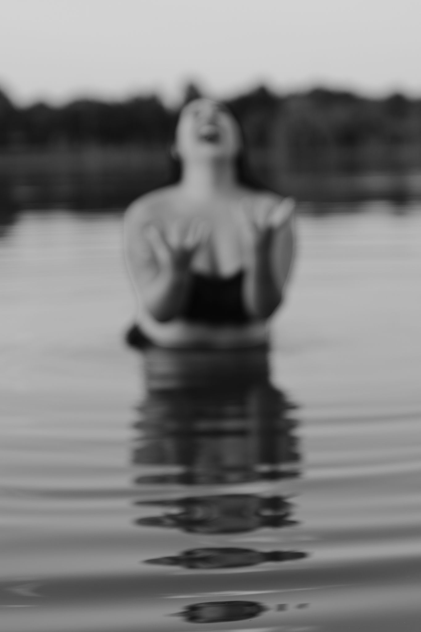 Black and white photo of a woman standing in a lake, head tilted back with hands raised, water reflecting her figure.