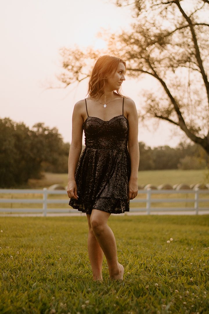 Young woman in black sequin dress standing barefoot on green grass at golden hour sunset near white fence.