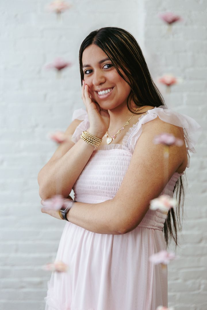 A person in a pink smocked dress poses against a white wall with floating pink flower petals.