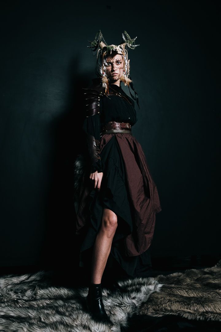 A person in a flowing black dress and dramatic fantasy-style crown poses against a dark backdrop in an artistic fashion shot.