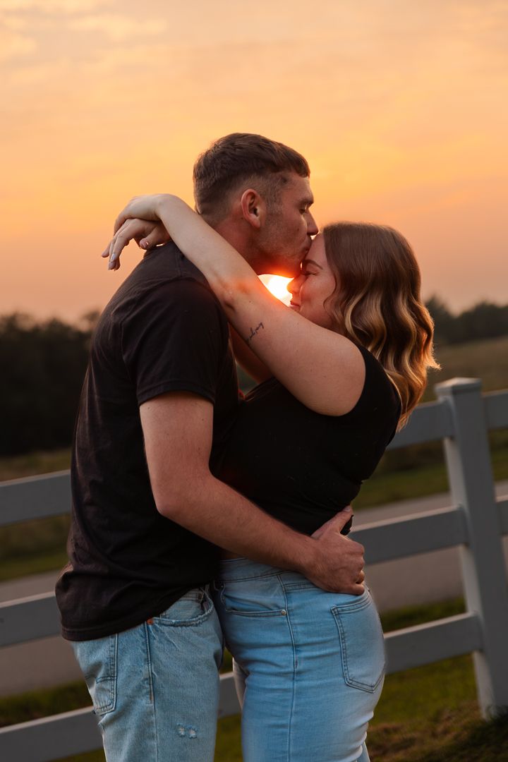 A couple embrace and kiss at sunset on a country road while leaning against a white fence.