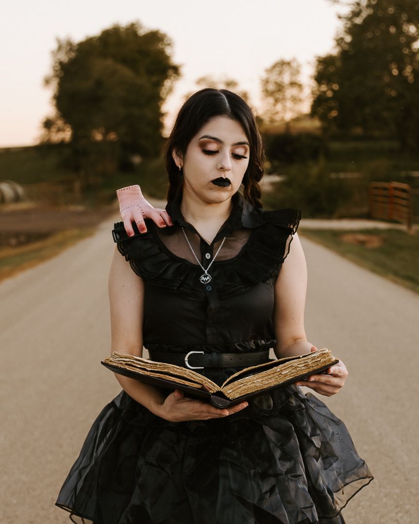 A person in a black gothic dress reads an antique book on a rural road at sunset.