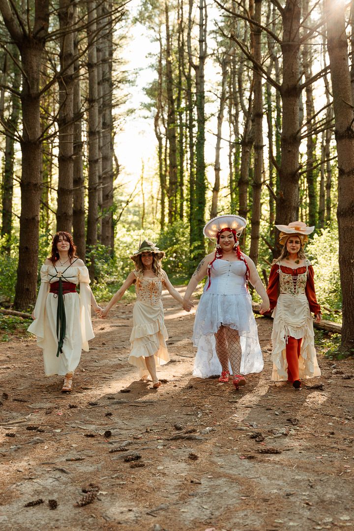 Four women in fantasy costumes walk through a sunlit forest path wearing medieval-inspired dresses and hats.