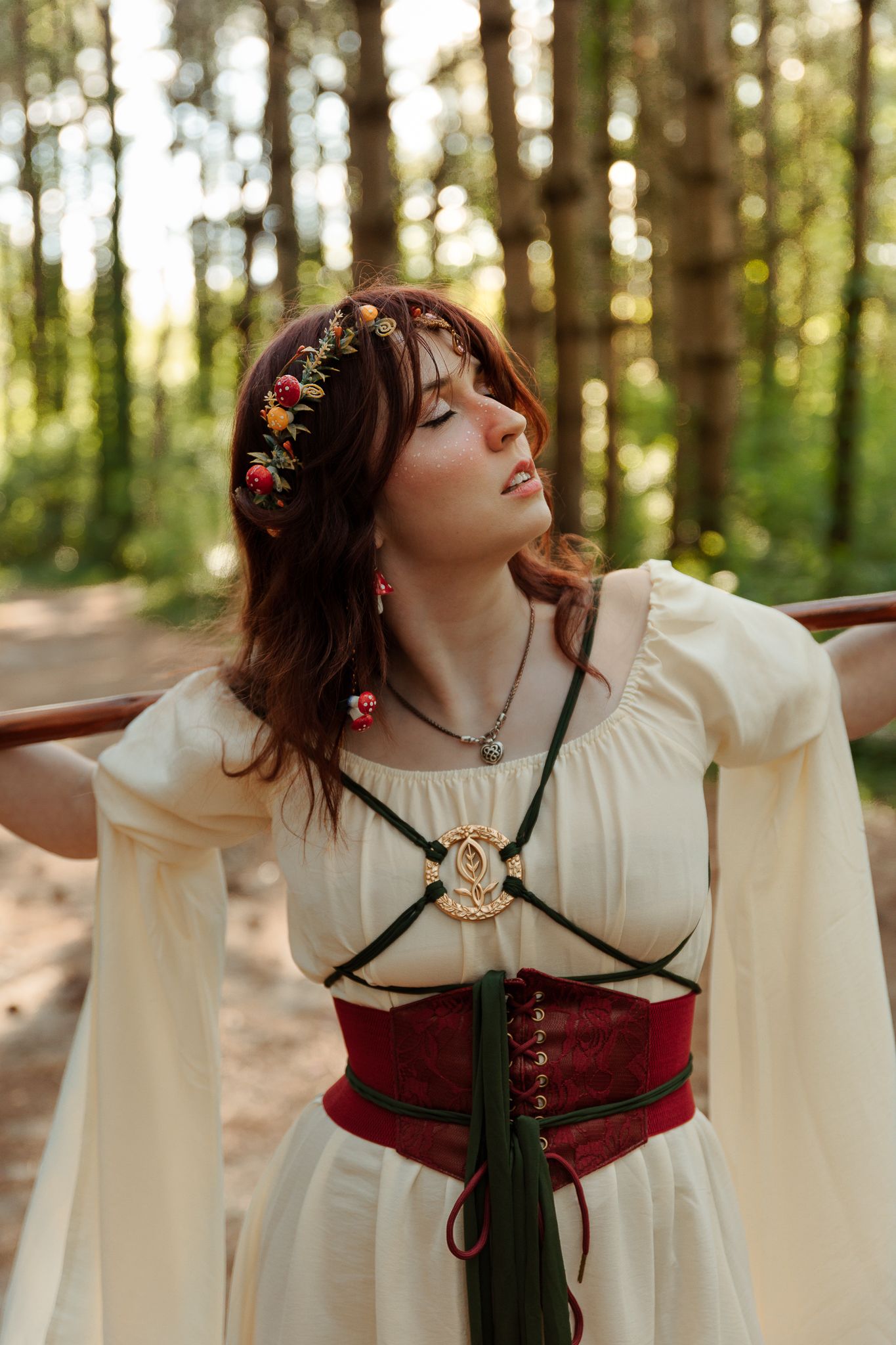 Woman in medieval fantasy dress with floral crown poses gracefully in a sunlit forest setting.