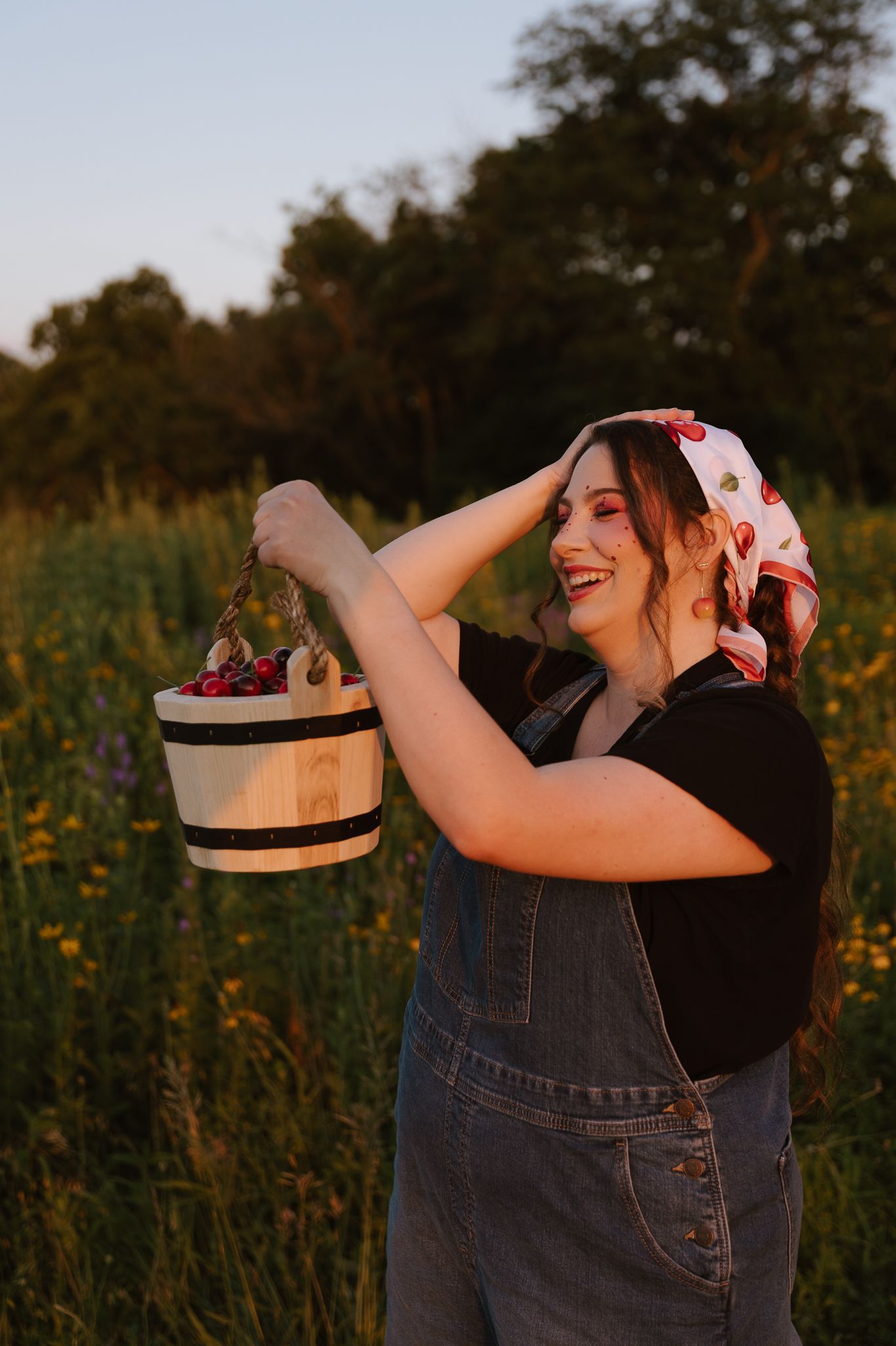 Smiling woman in denim overalls and cherry bandana holding a striped wooden basket of cherries in a sunlit field.