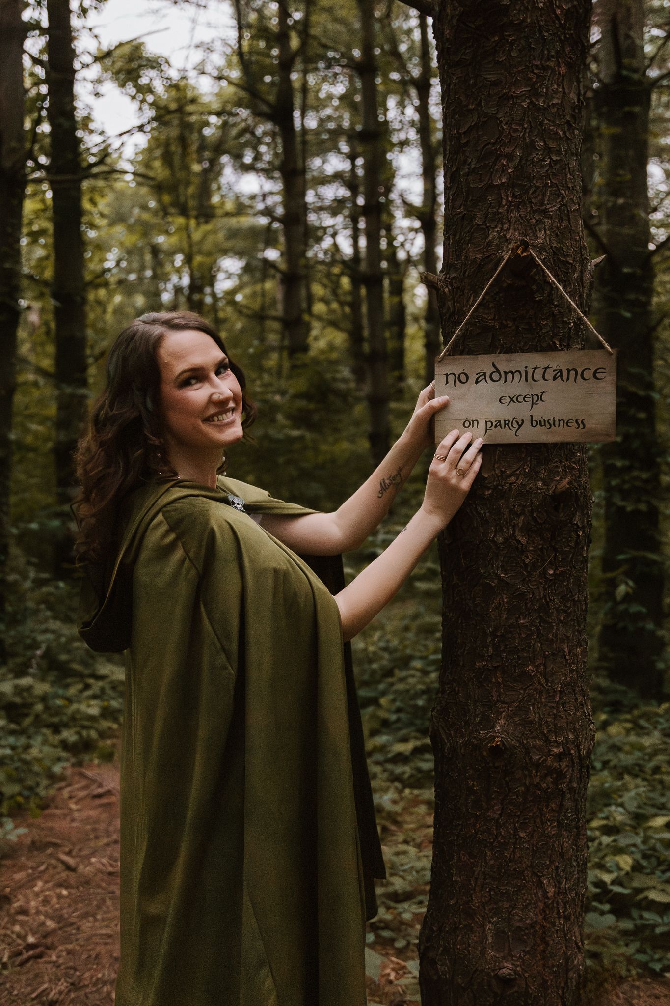 Smiling woman in green elvish cloak touching Lord of the Rings no admittance sign on forest tree.