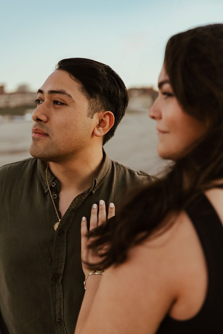 A couple stands near a waterfront at sunset, the woman showing off her engagement ring while the man gazes thoughtfully into the distance.