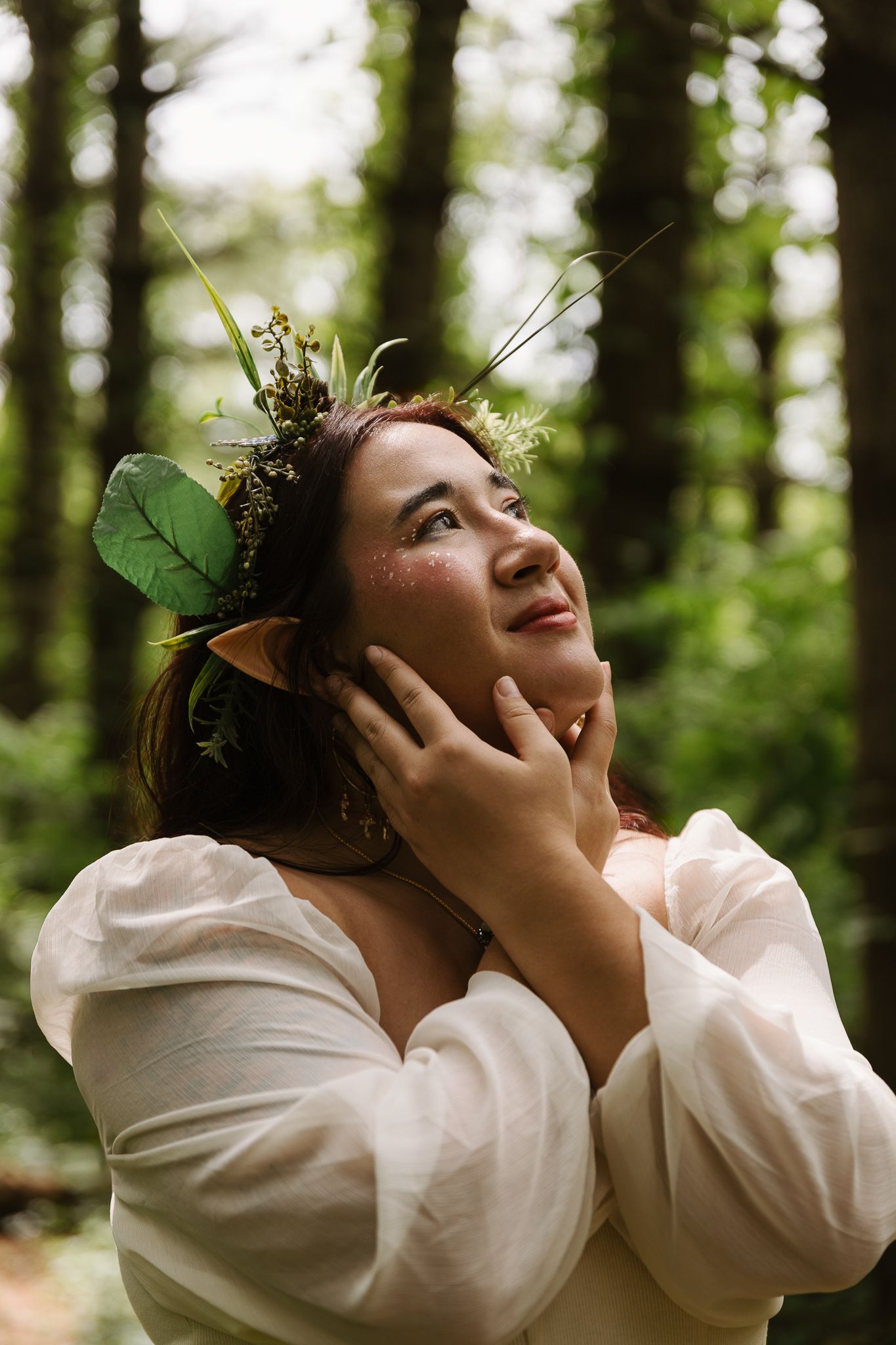 Woman in white dress with elf ears and floral crown poses in forest, looking up with hands framing her face.