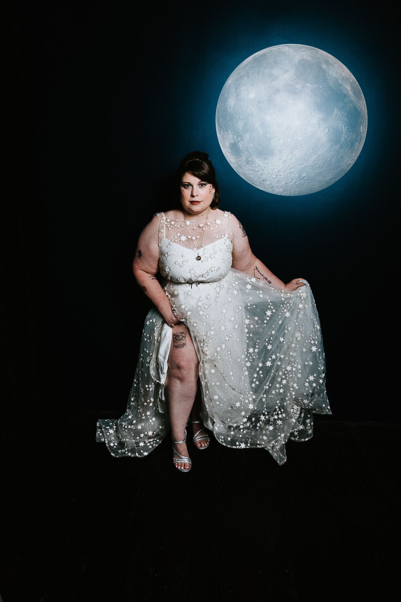 Plus-size woman in white starry gown poses elegantly against a dark background with a glowing full moon.