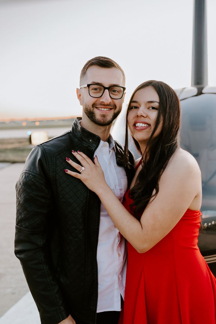 A couple in formal attire share a joyful moment together in front of a helicopter at dusk.