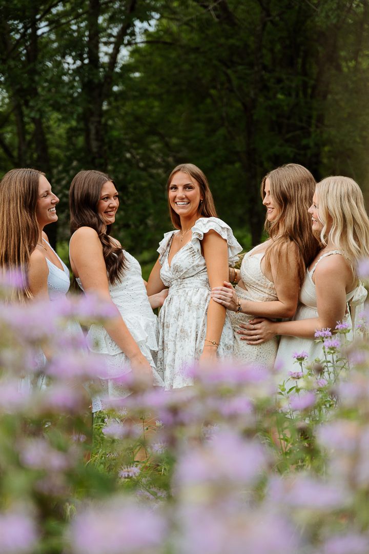 People in white dresses gather together among purple wildflowers in a sunny meadow.