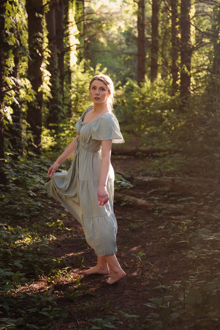 A woman in a vintage-style dress walks barefoot along a sunlit forest path.