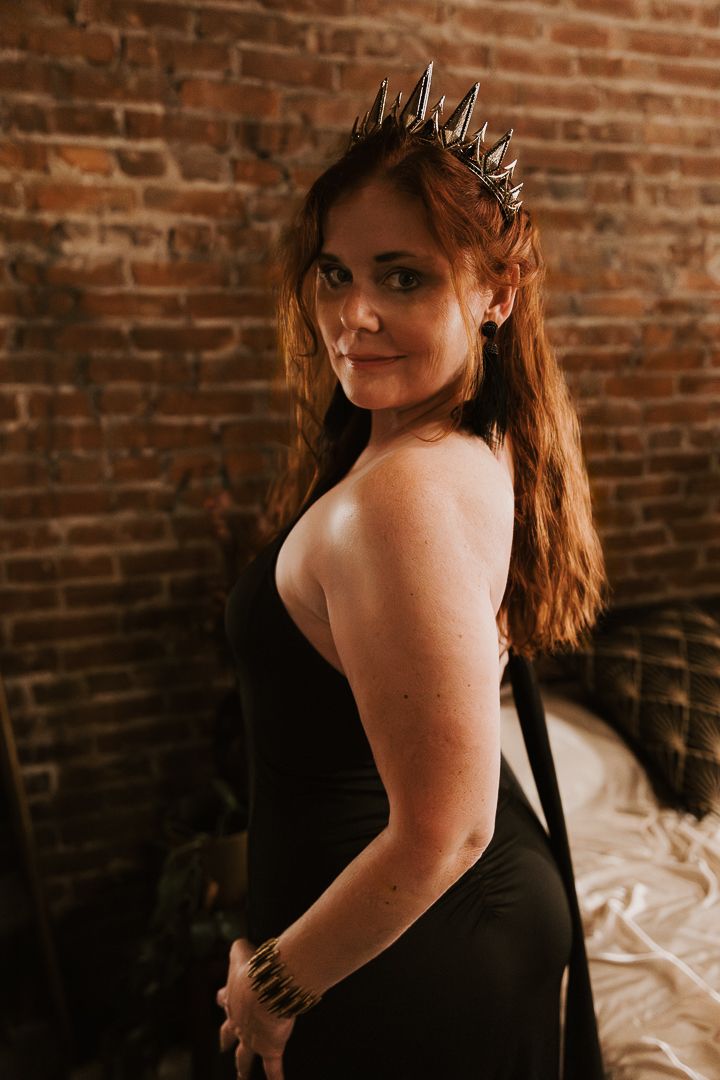 Woman in black dress poses against brick wall in moody boudoir photo series.