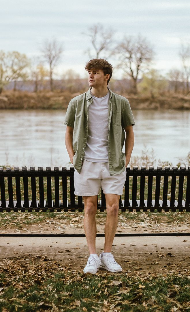 Someone rests on a black park bench surrounded by fallen leaves with a river in the background wearing casual attire.