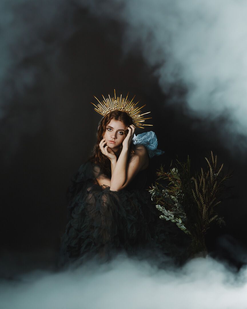 Ethereal portrait in dark misty setting with golden crown and blue dress against dramatic smoky background.