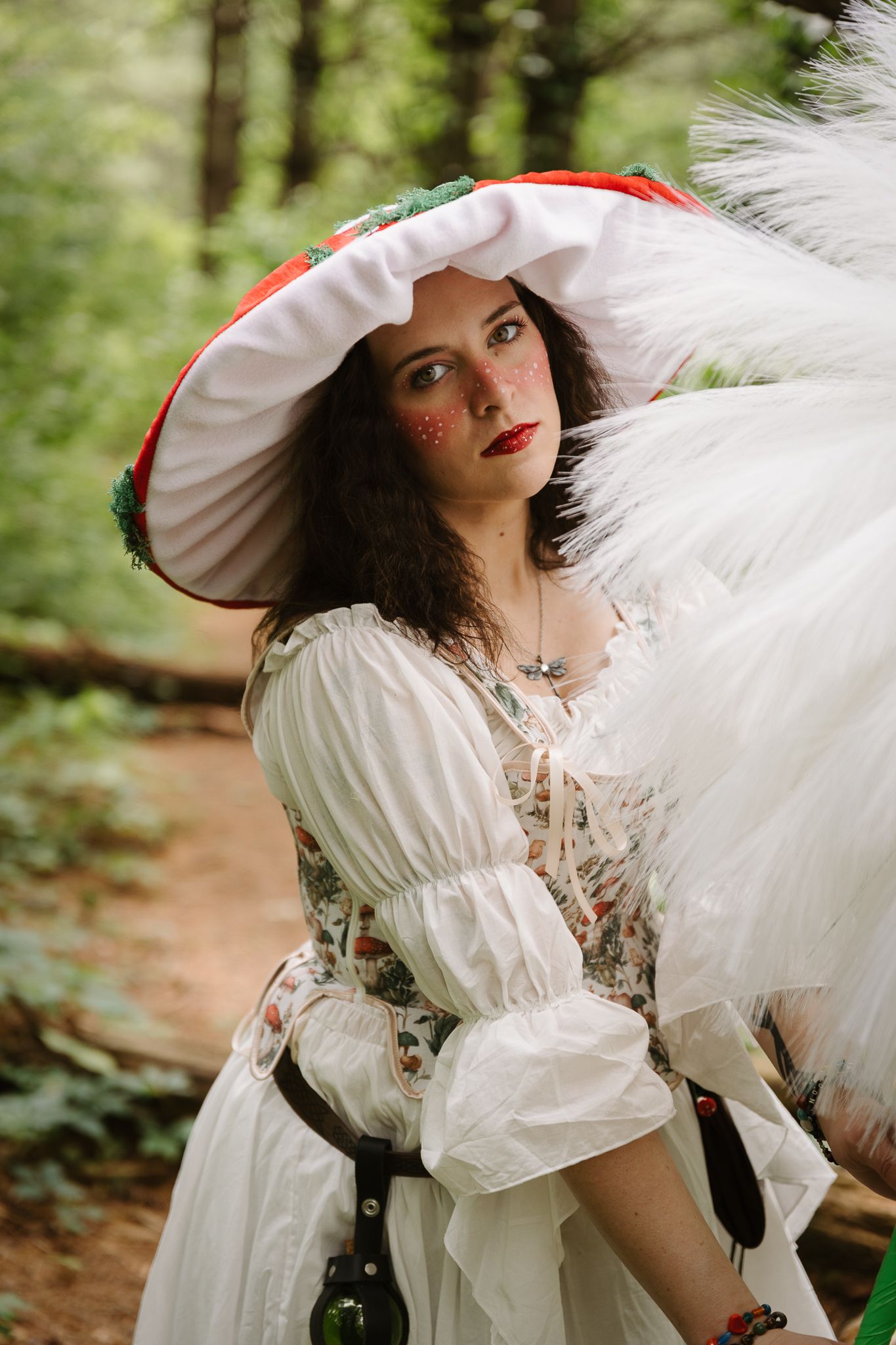 Woman in white floral dress and red mushroom hat poses in forest, wearing whimsical fairy tale costume with feathered accessories.