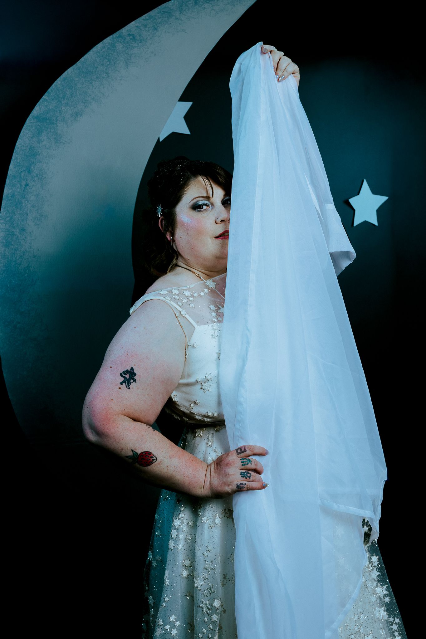 Woman in white wedding dress holding veil, posing before moon and stars backdrop with tattoos visible.