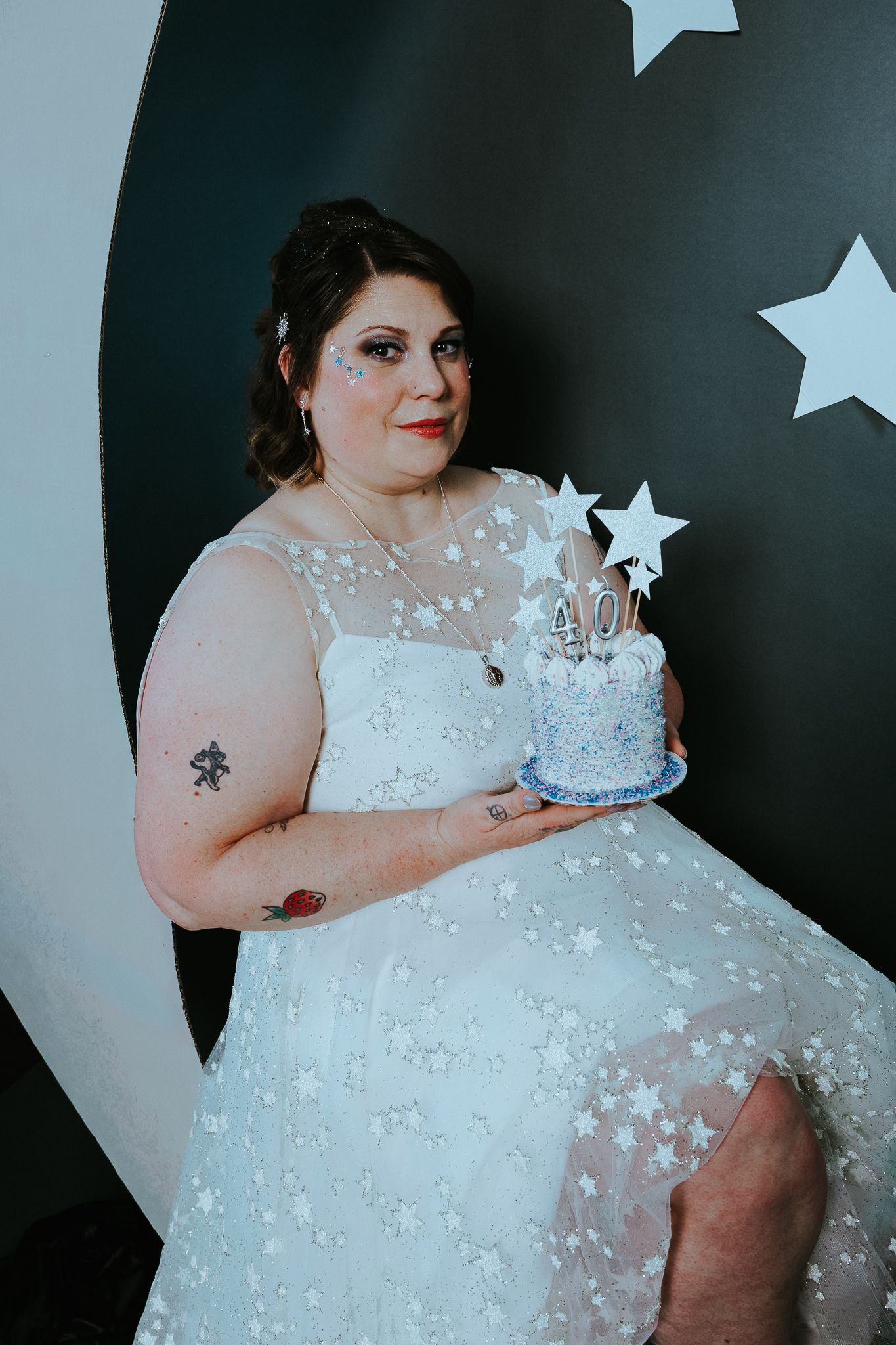 Plus-size bride in star-patterned white gown holds a blue star-topped wedding cake against a black backdrop.