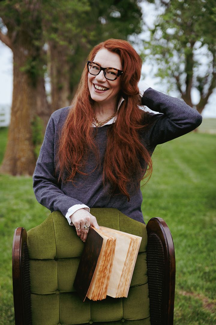 Person in black sweater holding an old book while seated in a plush green chair outdoors.