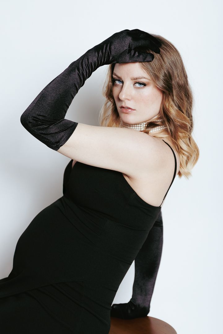 Fashion portrait of model in classic black dress and elbow-length gloves against white backdrop.