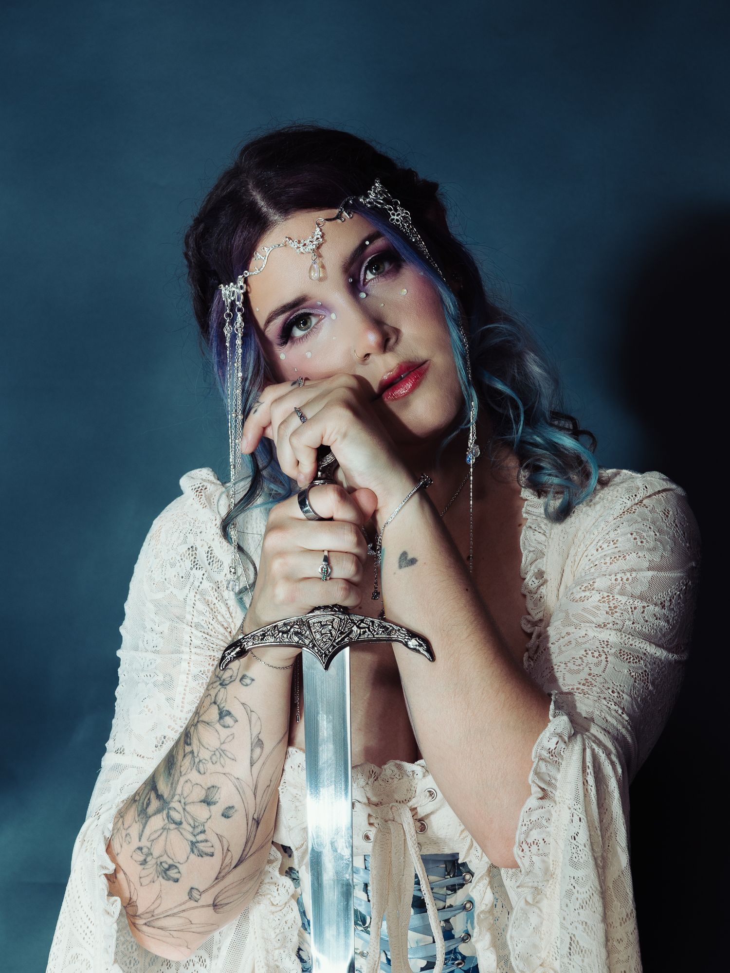 A mystical woman in white lace holds a silver sword, adorned with a jeweled headpiece and floral tattoos.
