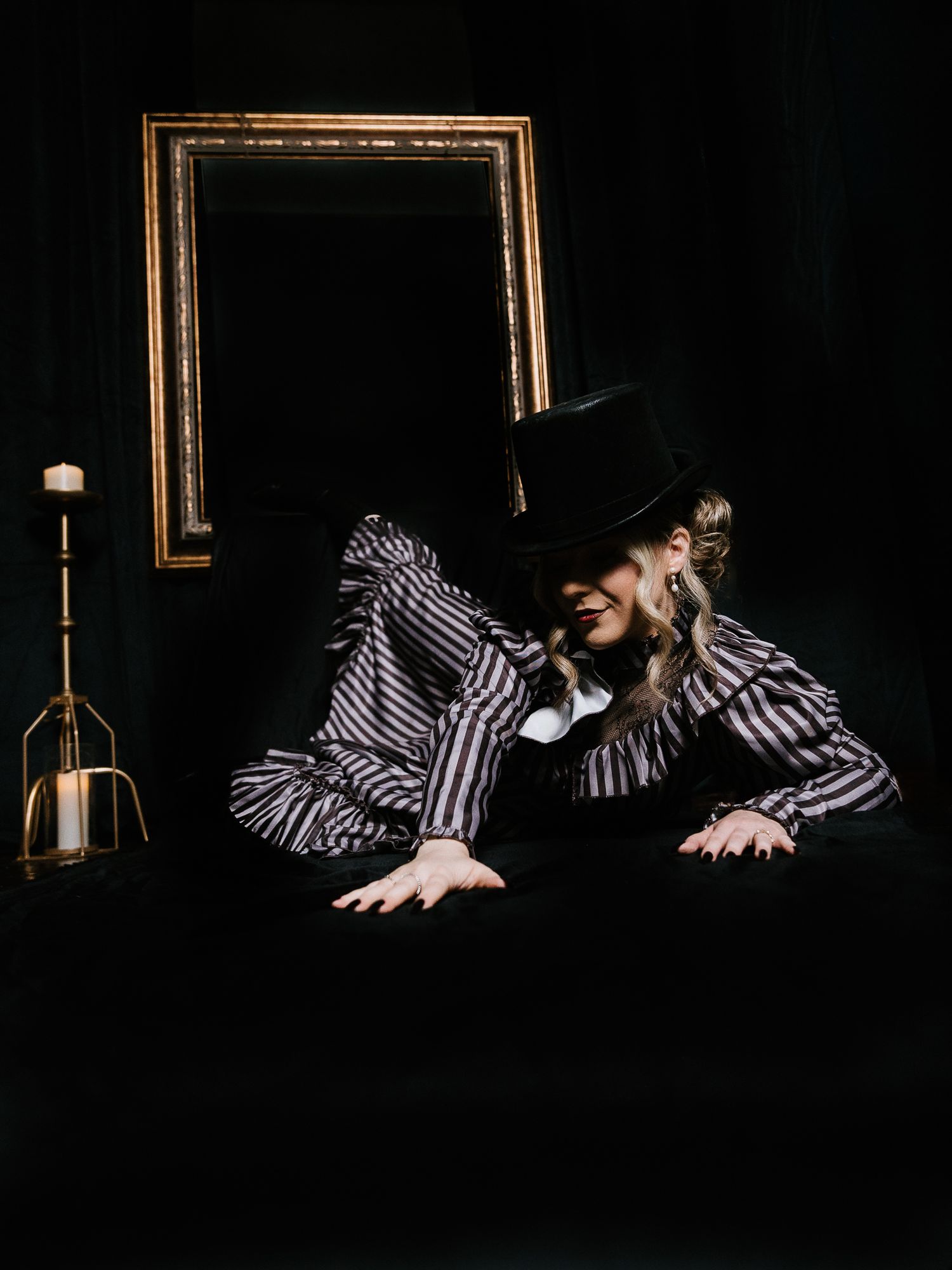 A mysterious performer in a striped blouse and black top hat leans over a dark surface beside a candle and ornate gold frame.