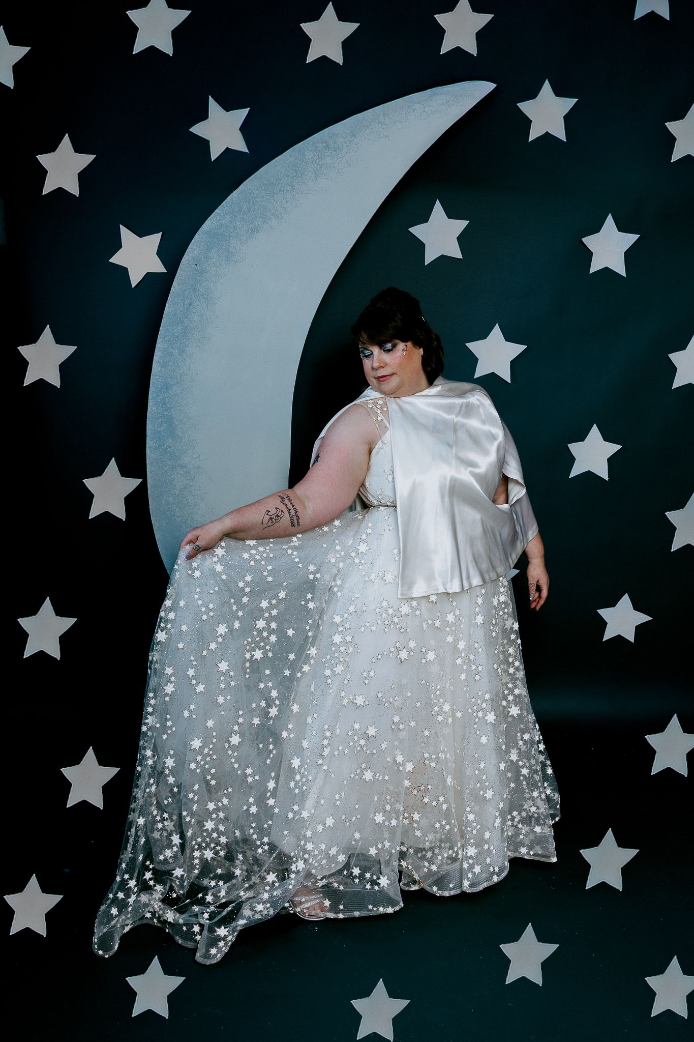 Plus-size bride in pearl-adorned gown leans against crescent moon prop with star-covered dark backdrop.