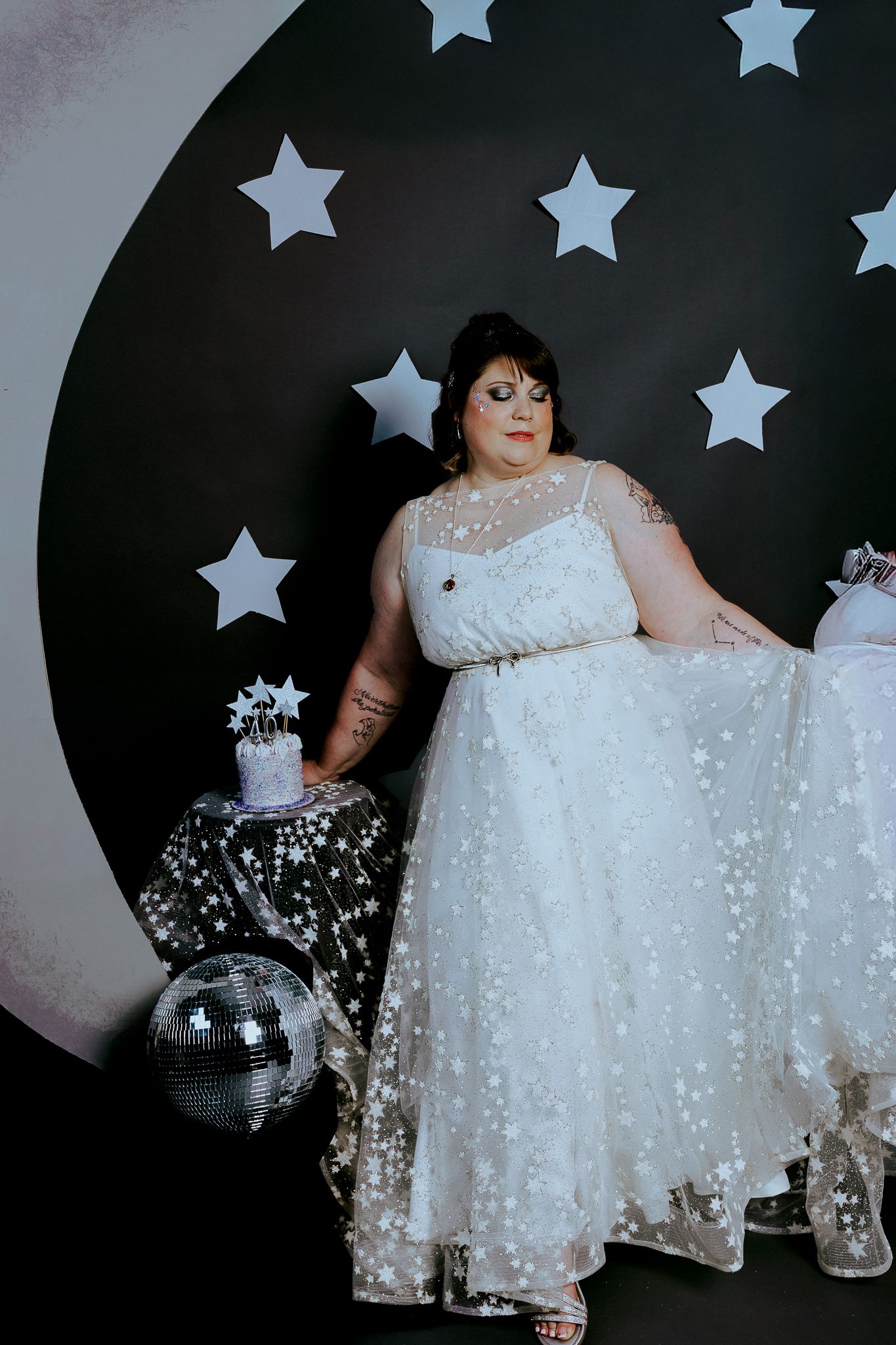 Plus-size bride in star-patterned white gown poses before black moon and stars backdrop with disco ball.