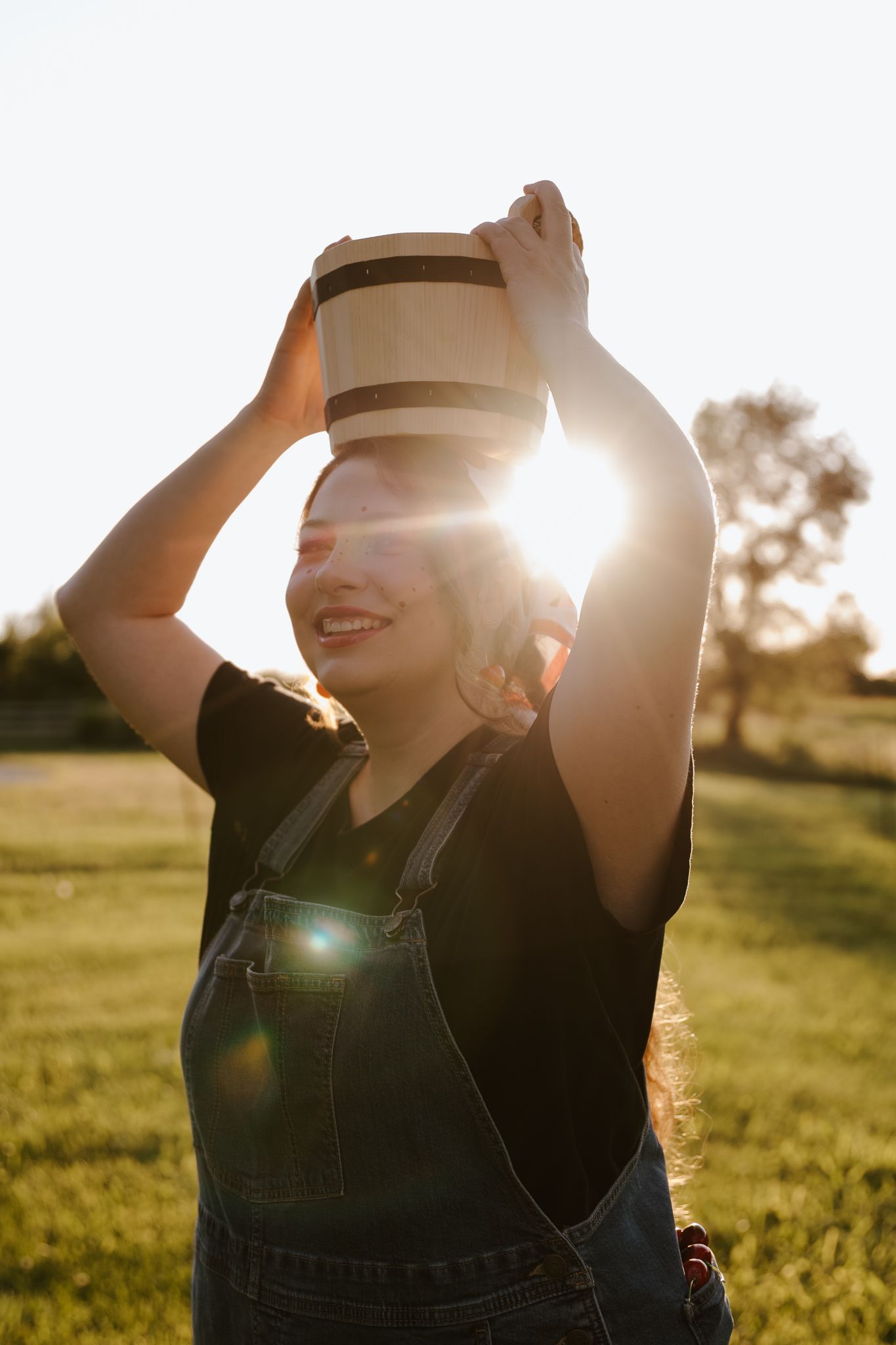 Woman in denim overalls balancing a wooden bucket on her head, smiling in a sunlit green field.