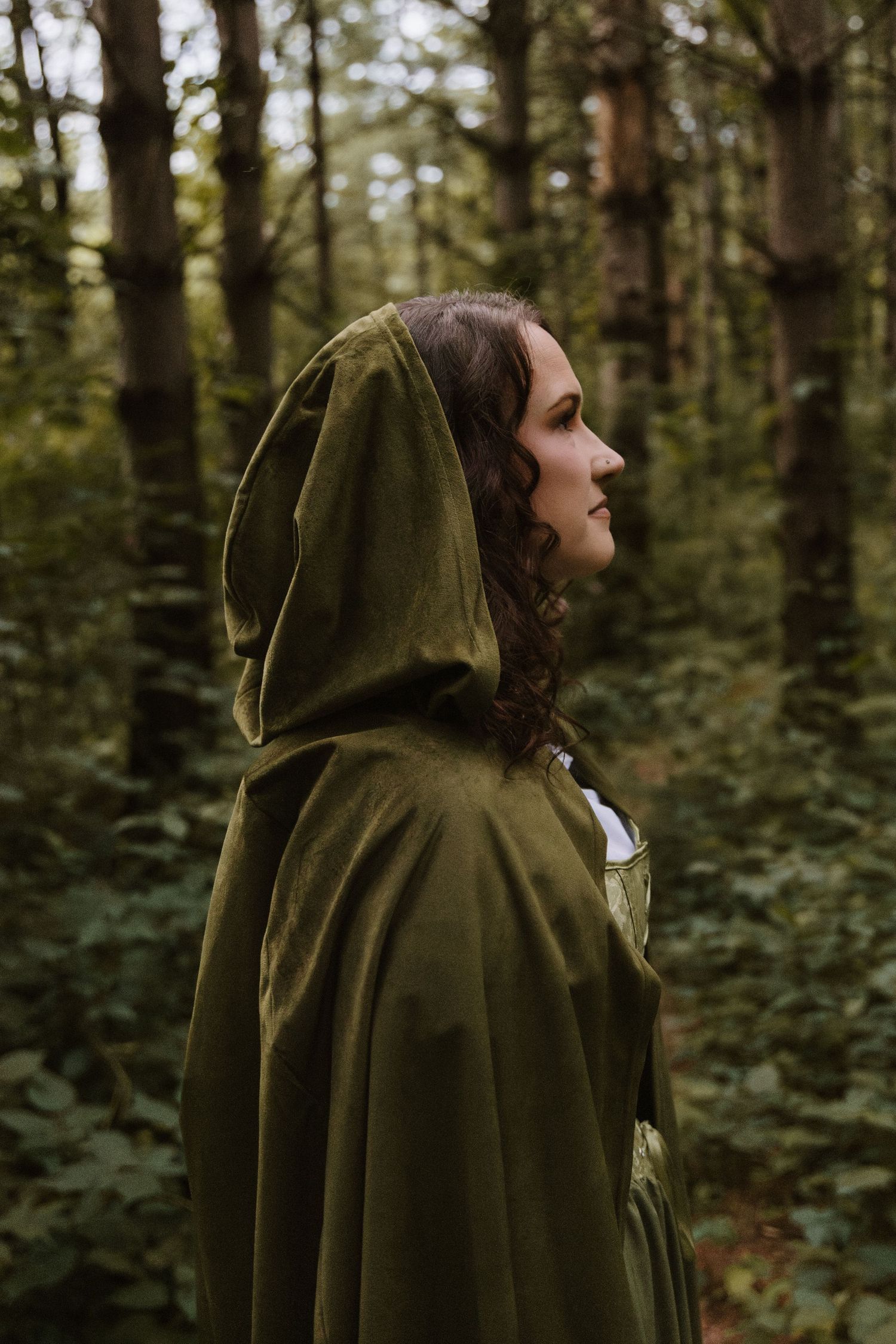 A woman in a green hooded cloak stands in a lush forest, gazing upward in a mystical, fairytale-inspired portrait.