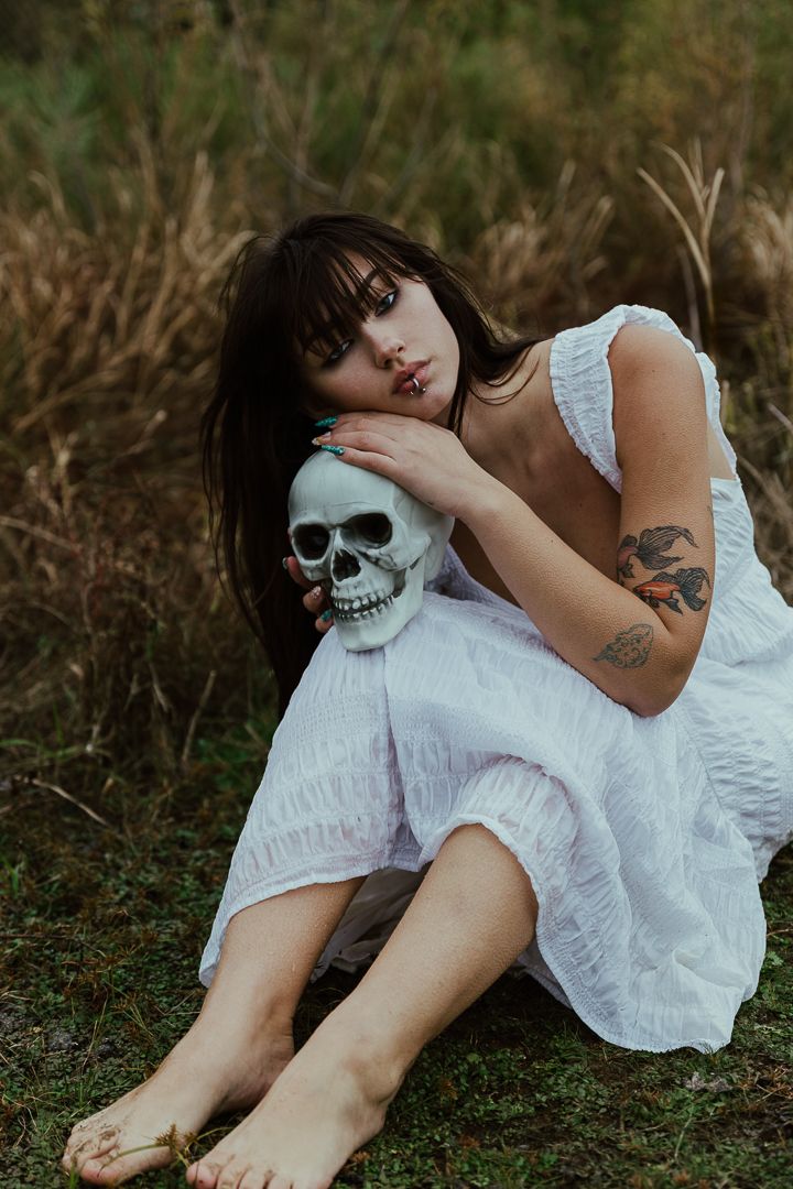 A person in a white dress lies on grass while wearing a skull mask in a haunting artistic photoshoot series.