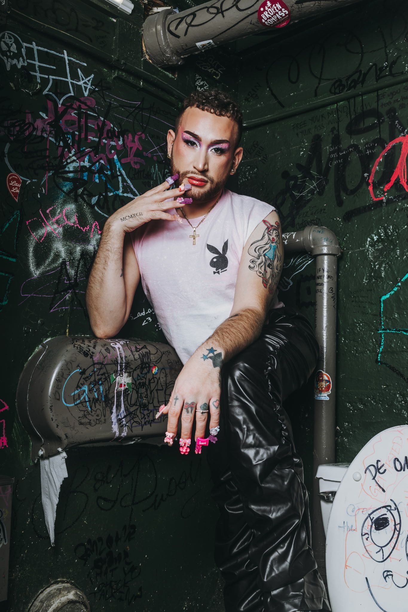 Person with tattoos and colorful nails poses against graffiti wall wearing Playboy tee and leather jacket.