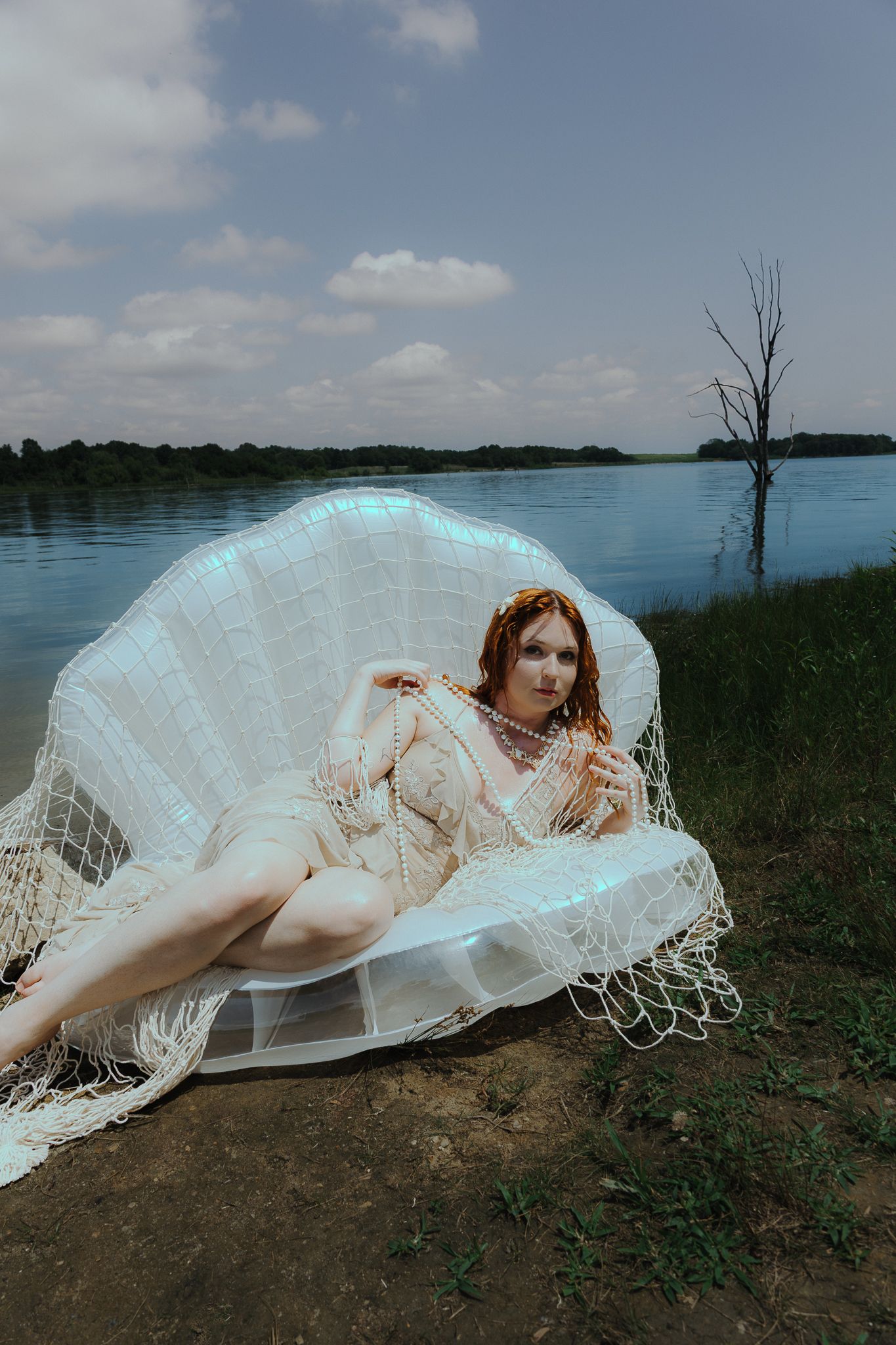 A woman in a lace dress reclines in a shell-shaped chair by a serene lake under a cloudy sky.