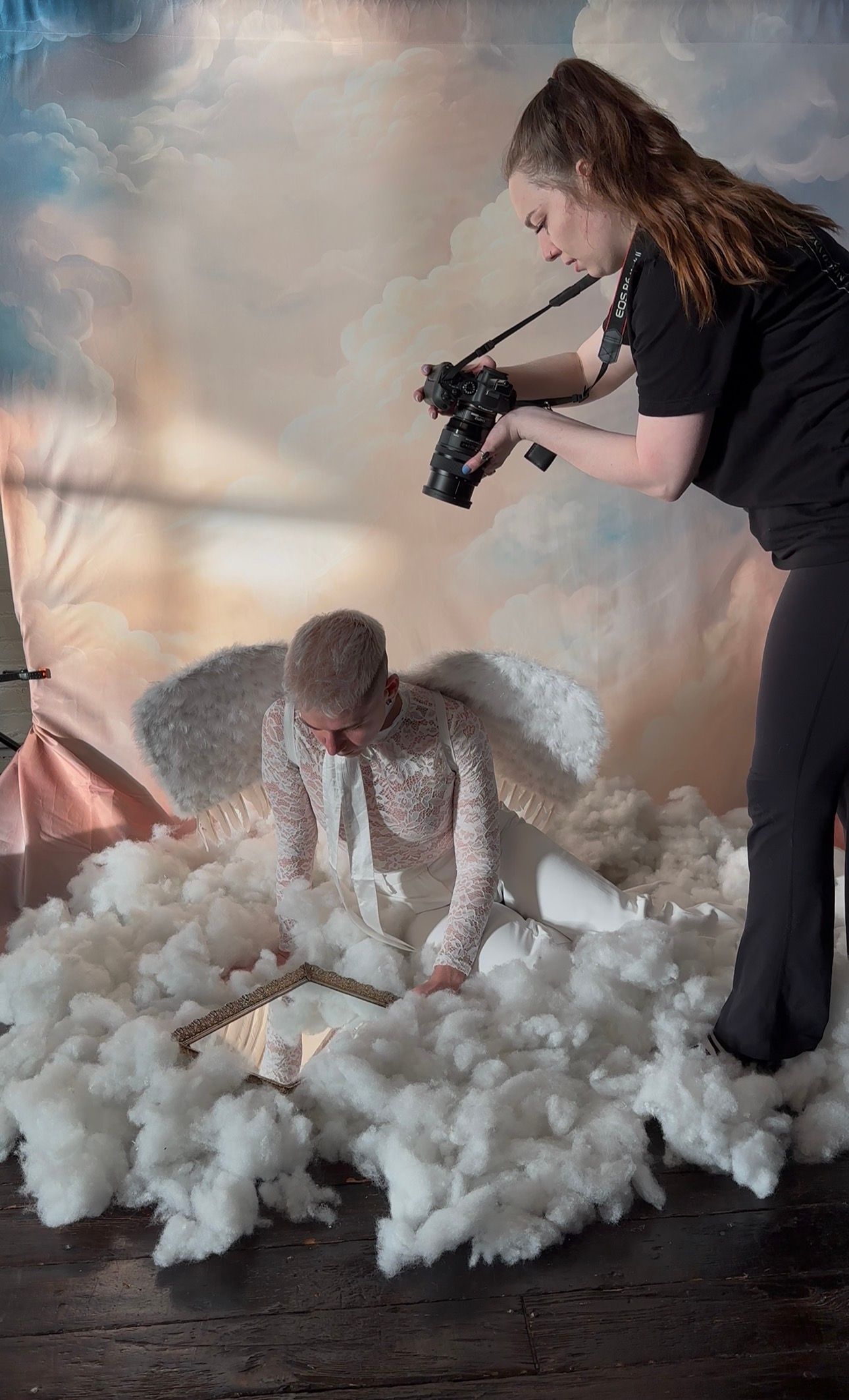 Photographer captures man dressed as angel with white wings amid fluffy clouds on a studio backdrop set.