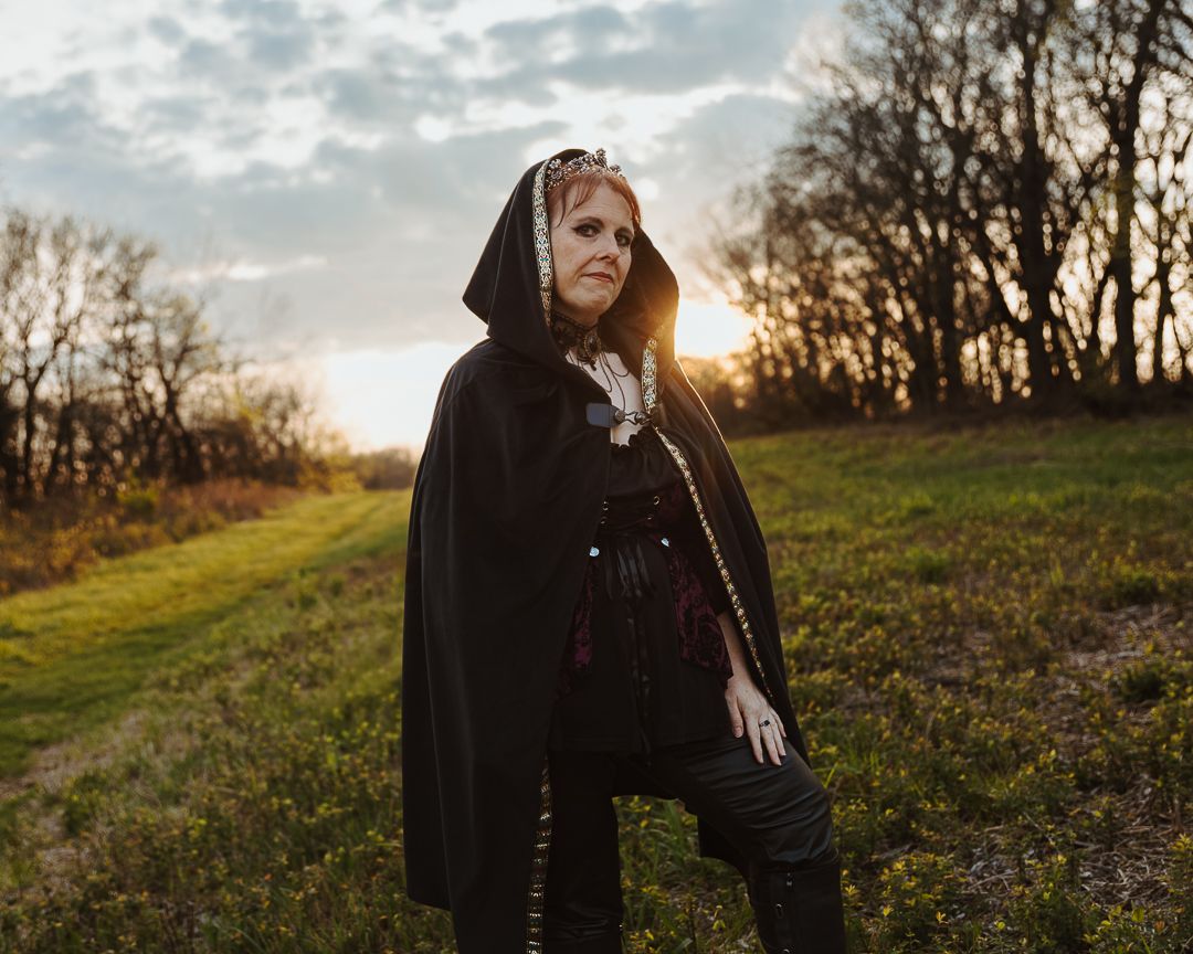 Woman in black medieval cape and hood standing in a field at golden hour sunset.