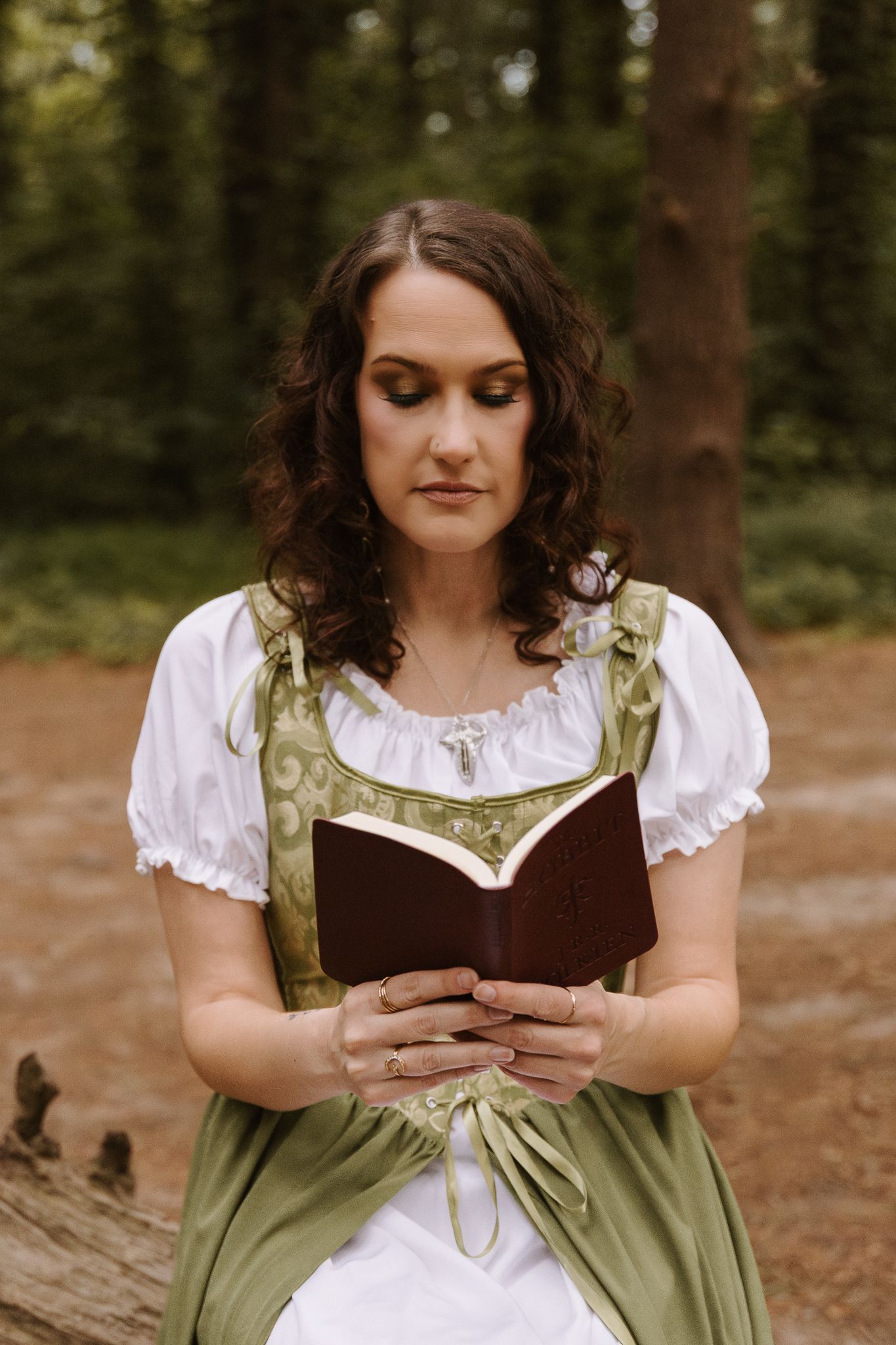 Woman in green and white medieval dress reads a brown leather journal in a forest setting.
