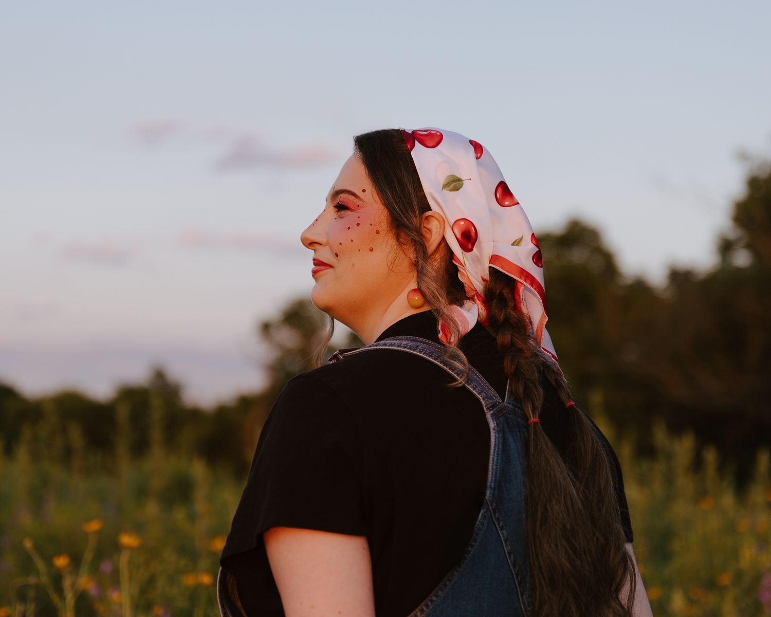 Young woman in cherry print headscarf and overalls standing in a field at golden hour sunset.