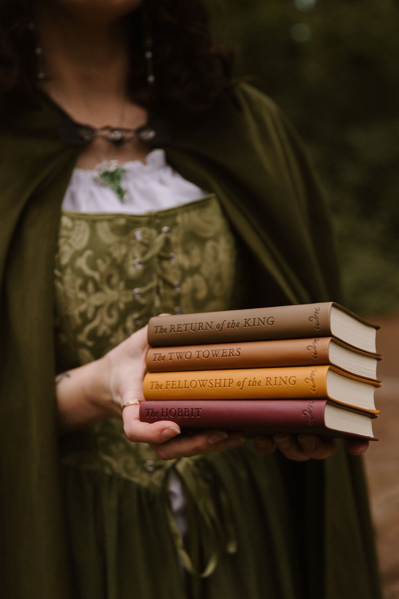 Woman in green medieval dress holding stack of Tolkien books including The Hobbit and LOTR trilogy.