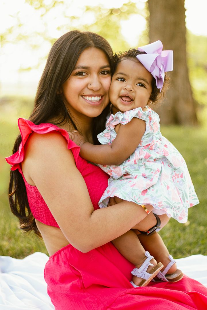 Two people wearing pink and white outfits embrace joyfully while sitting on grass during an outdoor photoshoot.