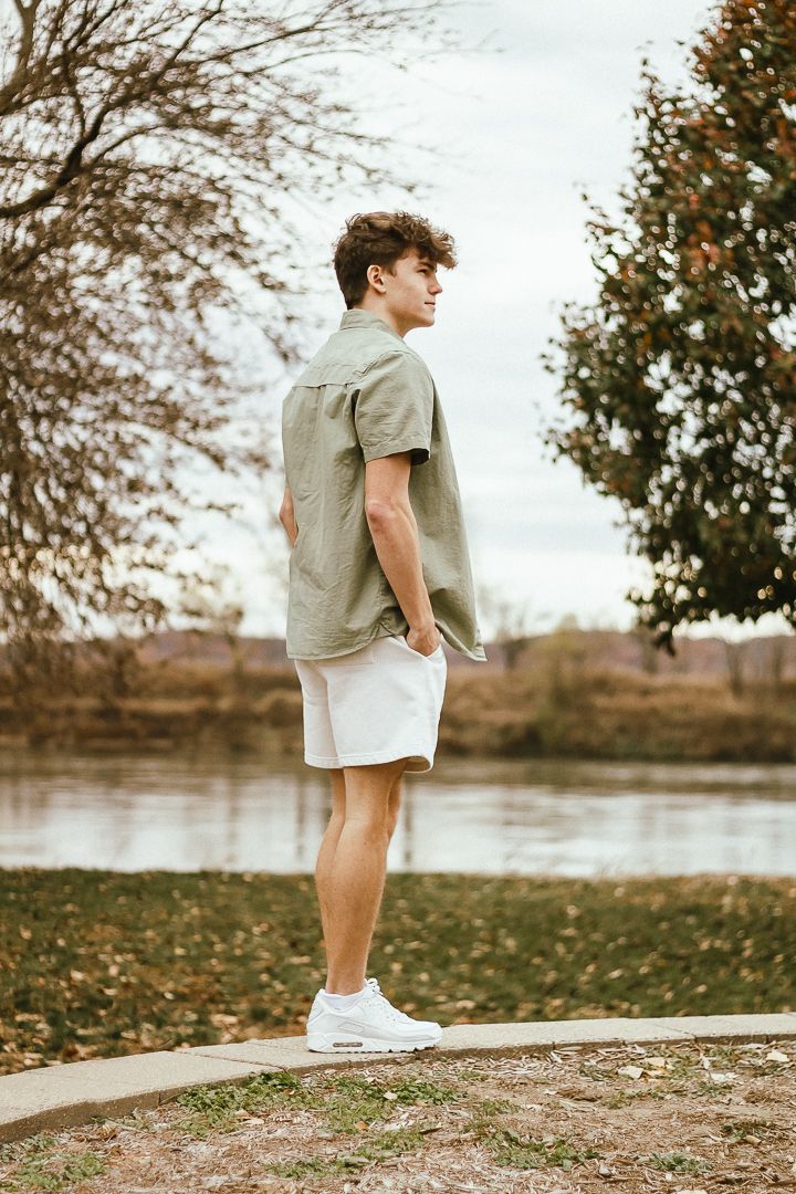 Someone stands in casual summer attire by a lake on an autumn day with fallen leaves and trees in the background.