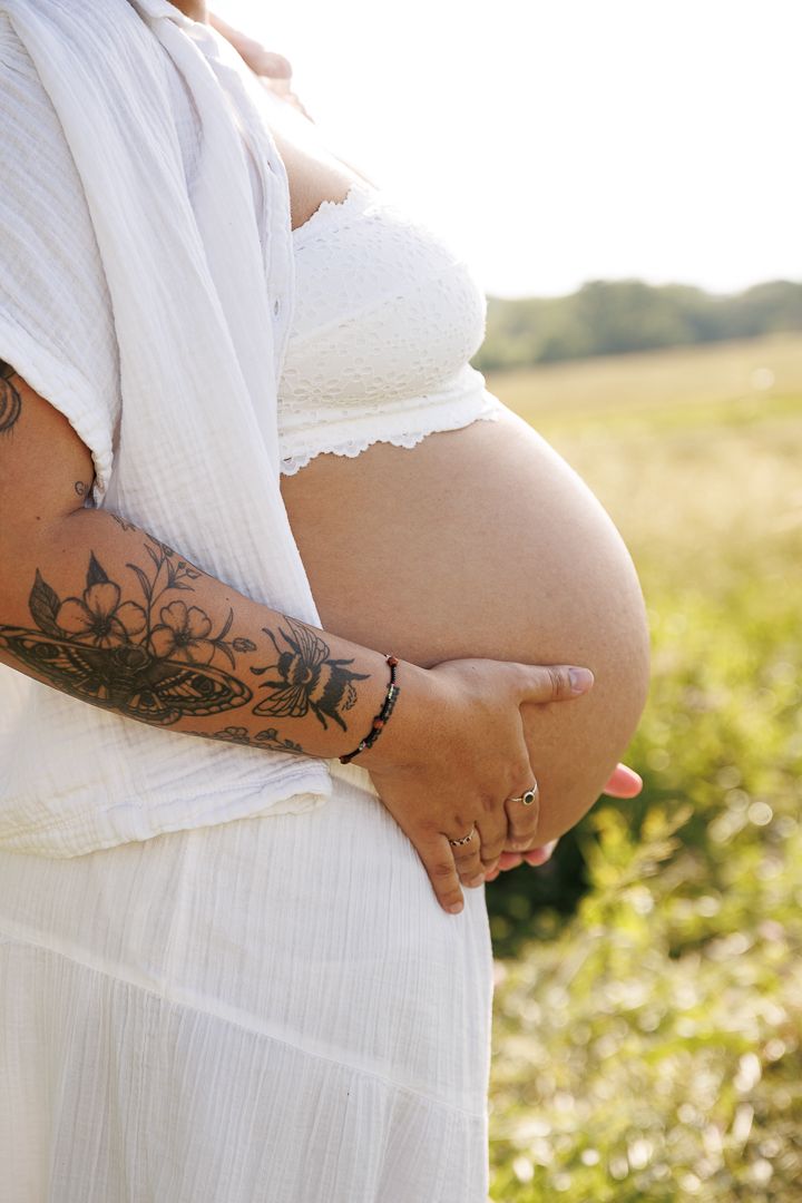 Close up shots of pregnant belly with visible arm tattoos in a flowing white maternity dress.
