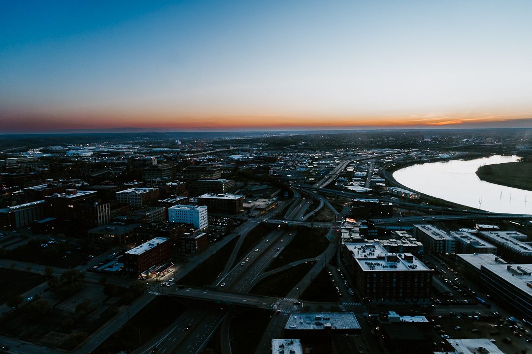Aerial cityscape view showing urban streets and a curving river at dusk with orange and blue skies.