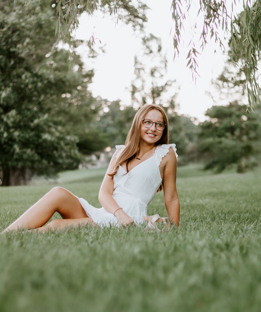 A woman in a white lace dress poses gracefully on lush green grass in a dreamy outdoor photoshoot setting.