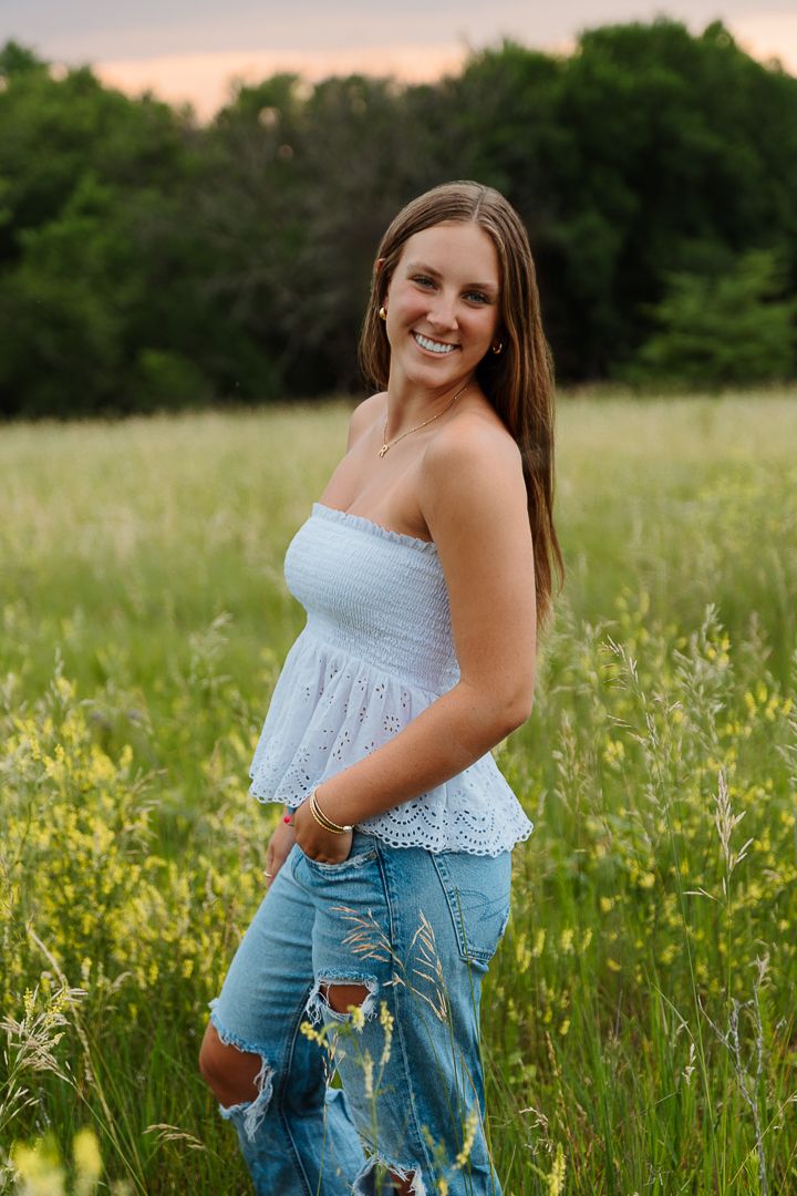 Person in light blue top and distressed denim poses in a wildflower meadow at sunset.