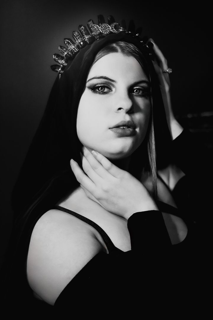 Black and white gothic portrait of a woman wearing a dark veil and a spiked crystal crown, with her hand touching her neck.