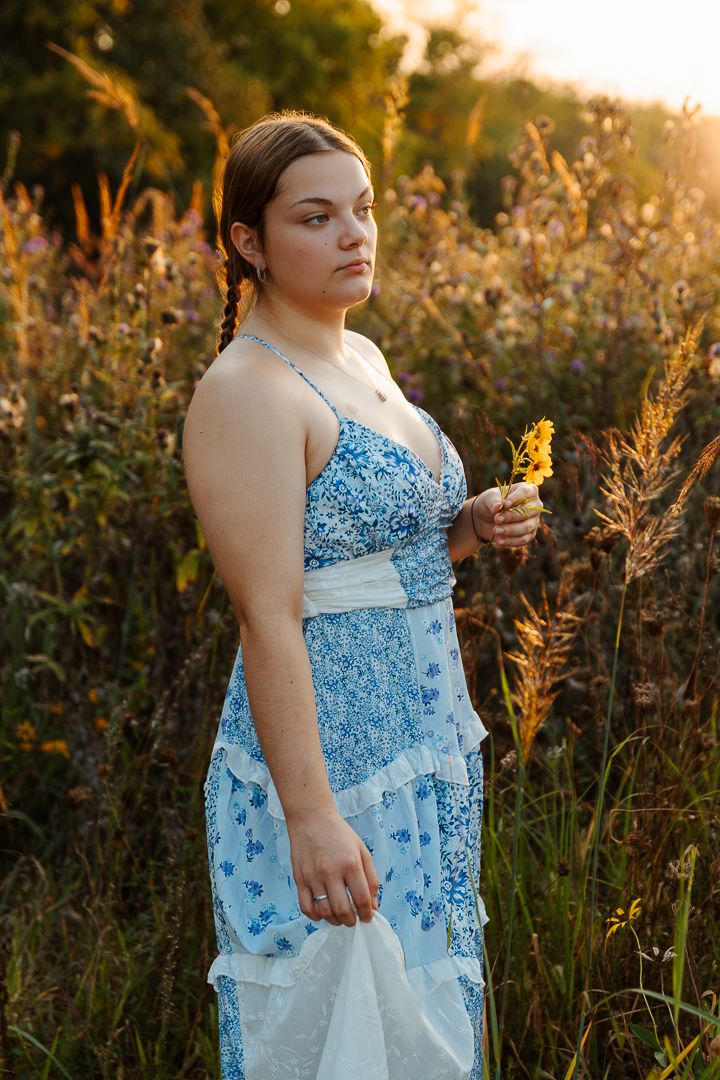 A person in a light blue floral dress stands among tall wildflowers during golden hour sunset.