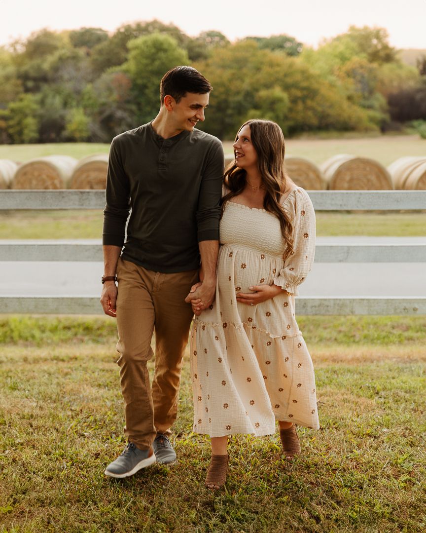 A couple walk together across a grassy field at sunset near hay bales, capturing a romantic rural moment.