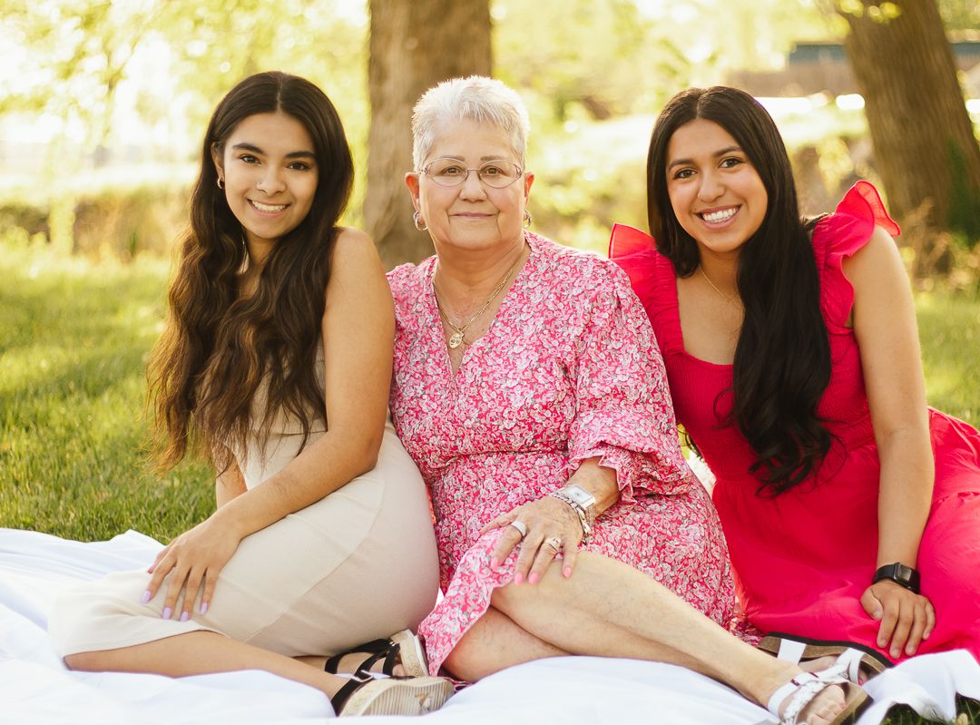 Three generations of family members wearing pink and white outfits sit together on grass during a sunny outdoor photoshoot.