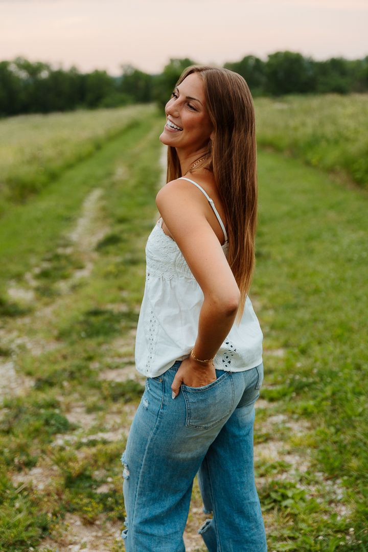 Woman in white top and jeans poses candidly walking through a sunny field with a dirt path.
