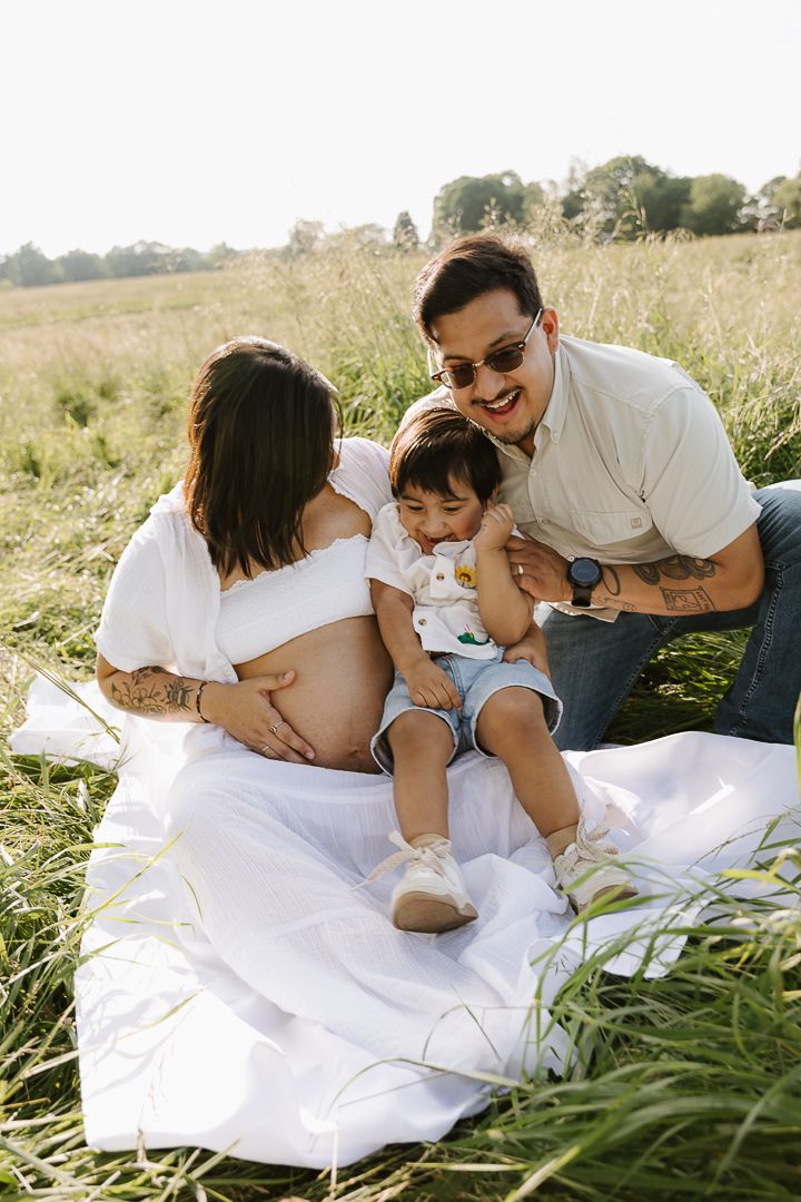 Family maternity photoshoot on a white blanket in a grassy field during golden hour.